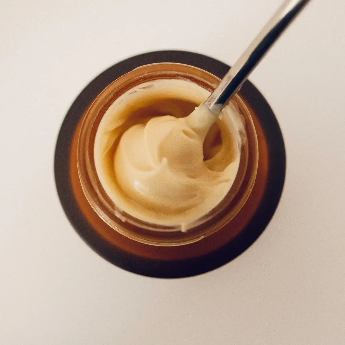 A creamy beige skincare product in a dark amber glass jar with a black lid, featuring a silver spoon resting inside.